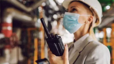 Mujer con casco y mascarilla usando radio en una fábrica.