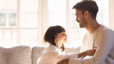 Niña jugando a ser doctora revisa a su padre con un estetoscopio en casa.