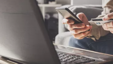 Persona comprando en línea desde un sofá, usando teléfono y tarjeta de crédito.