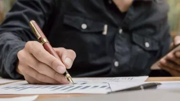 Persona escribiendo con bolígrafo sobre gráficos en papel, sosteniendo un smartphone.