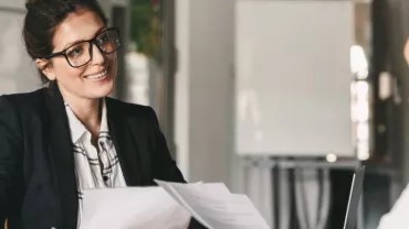 Mujer con gafas sonriendo mientras sostiene documentos en una reunión.