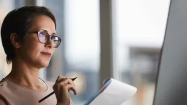 Mujer con gafas mirando el monitor mientras toma notas.