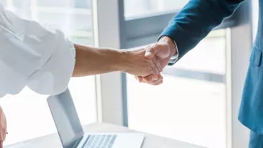 Dos personas estrechando manos sobre un escritorio con una computadora portátil.