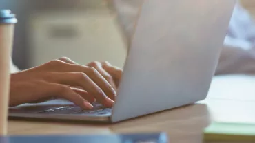 Persona escribiendo en un portátil con una taza de café al lado.