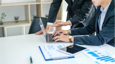 Dos personas en traje trabajando en una laptop con gráficos sobre la mesa.