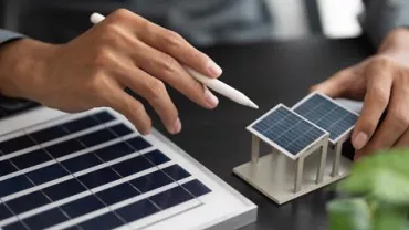 Manos sosteniendo un bolígrafo y un modelo de panel solar en miniatura.