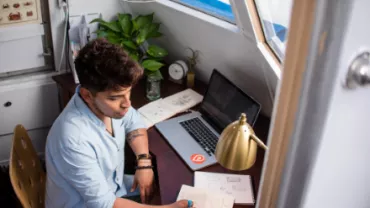 Persona escribiendo en una mesa con portátil y planta.