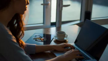 Mujer trabajando en computadora portátil cerca de una ventana con vista al agua.