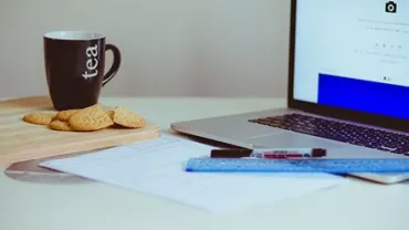 Taza de té junto a galletas, cuaderno, laptop y regla en un escritorio.