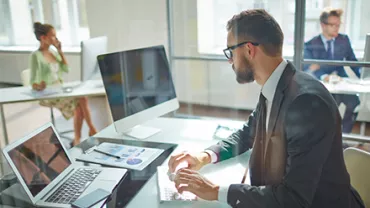 Hombre con gafas trabajando en una oficina moderna con computadora y gráficos.