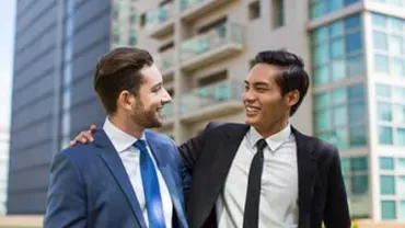Dos hombres de negocios sonrientes, vestidos con traje y corbata, hablando afuera de un edificio moderno.