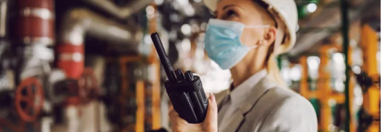 Mujer con casco y mascarilla usando radio en una fábrica.
