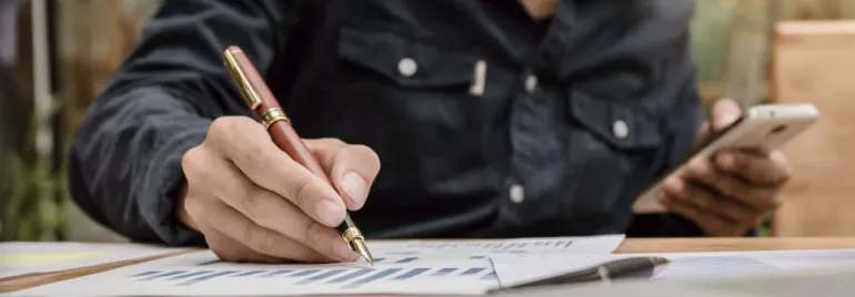 Persona escribiendo con bolígrafo sobre gráficos en papel, sosteniendo un smartphone.