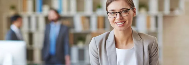 Mujer sonriente con gafas en una oficina con libros y personas al fondo.