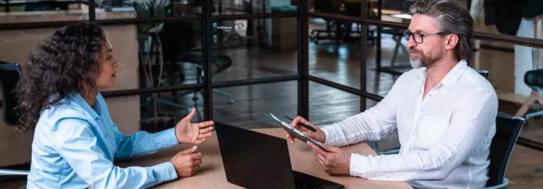 Dos personas conversando en una oficina, una con portátil y otra con tableta.
