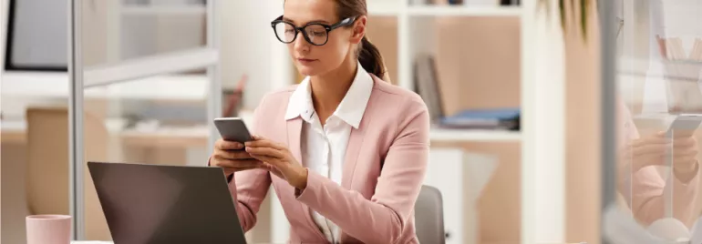 Mujer en oficina usando teléfono y portátil.