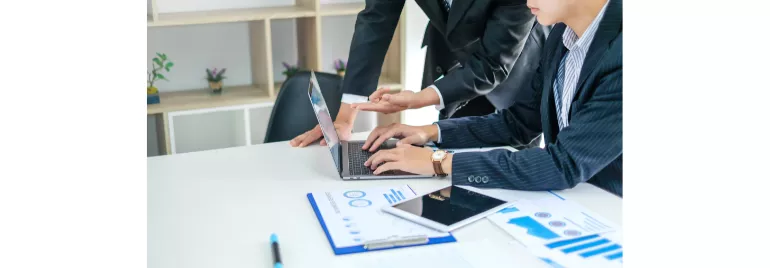 Dos personas en traje trabajando en una laptop con gráficos sobre la mesa.