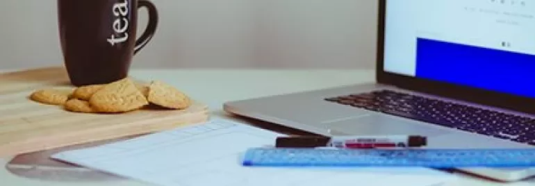 Taza de té junto a galletas, cuaderno, laptop y regla en un escritorio.