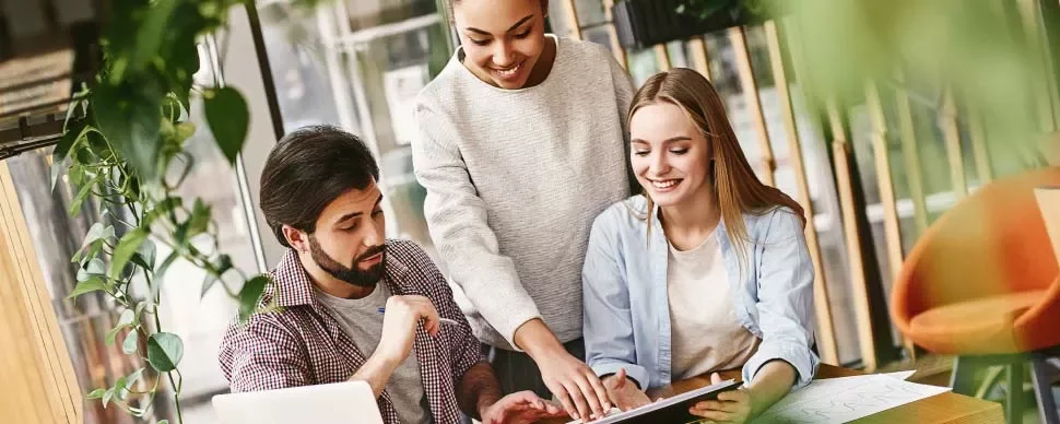 Tres personas colaborando en un proyecto en una mesa, rodeados de plantas.