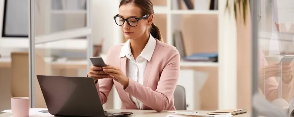 Mujer en oficina usando teléfono y portátil.