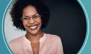 Mujer sonriente con gafas y cabello rizado, vestida con blusa rosa.
