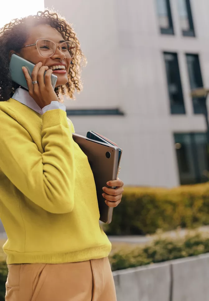 Professional speaking on a mobile phone while carrying a laptop outdoors in a modern business district, representing connectivity, flexibility, and client-focused professional services.