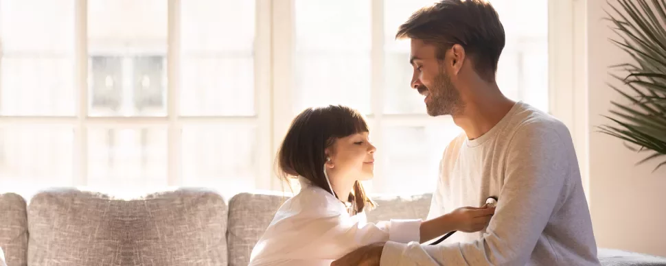 Niña jugando a ser doctora revisa a su padre con un estetoscopio en casa.