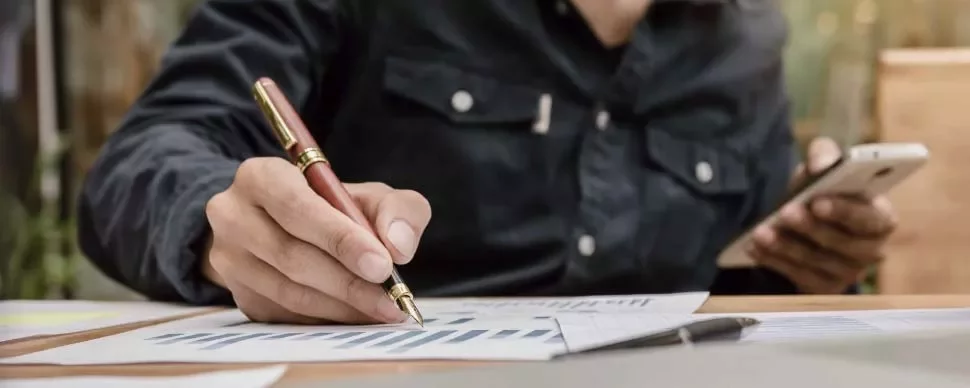 Persona escribiendo con bolígrafo sobre gráficos en papel, sosteniendo un smartphone.