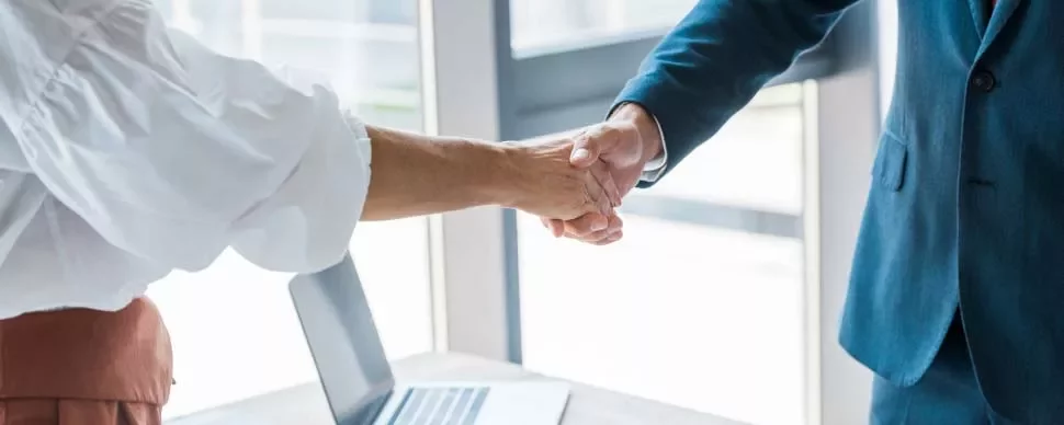 Dos personas estrechando manos sobre un escritorio con una computadora portátil.