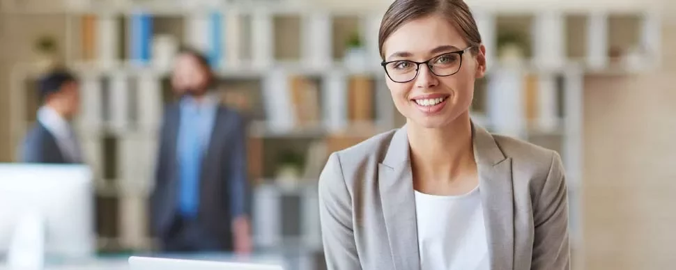 Mujer sonriente con gafas en una oficina con libros y personas al fondo.