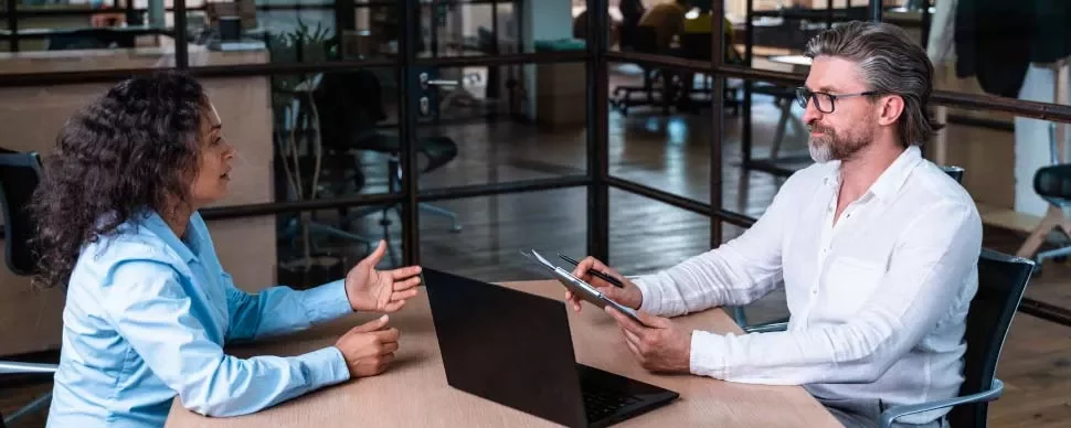 Dos personas conversando en una oficina, una con portátil y otra con tableta.