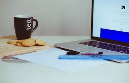 Taza de té junto a galletas, cuaderno, laptop y regla en un escritorio.