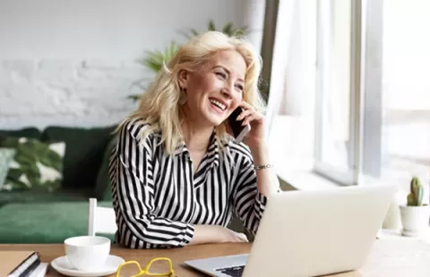Mujer sonriendo hablando por teléfono junto a computadora.